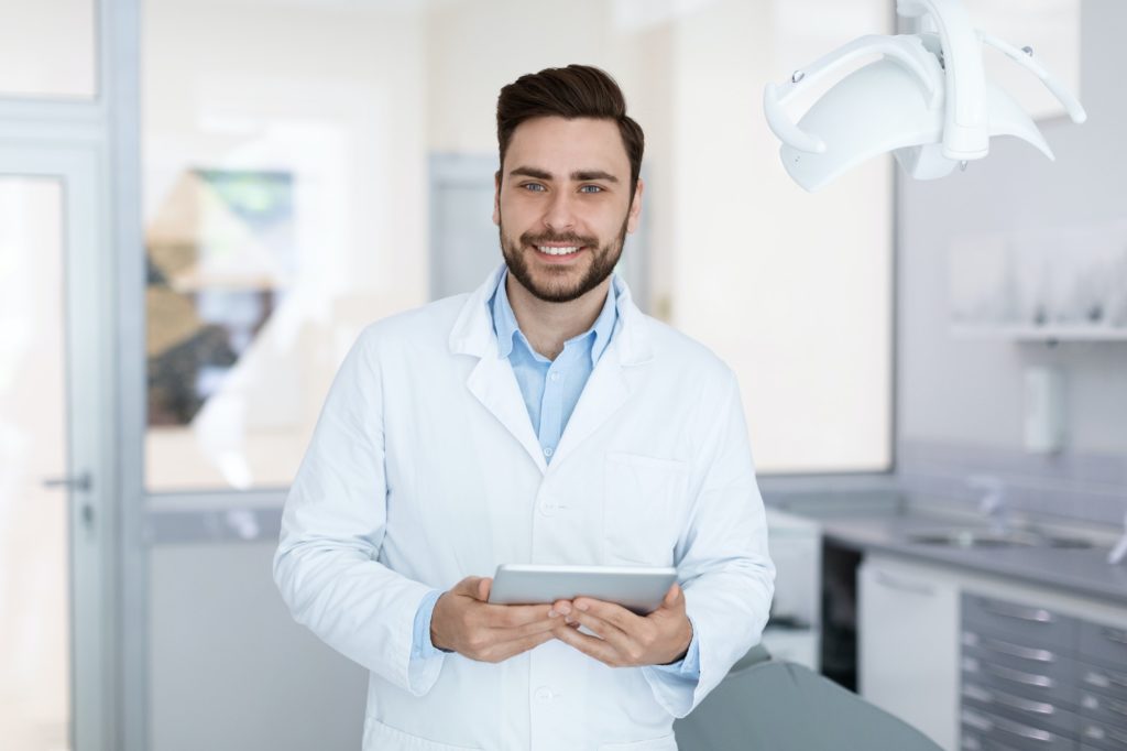 Delighted dentist smiling and holding digital tablet in dental cabinet